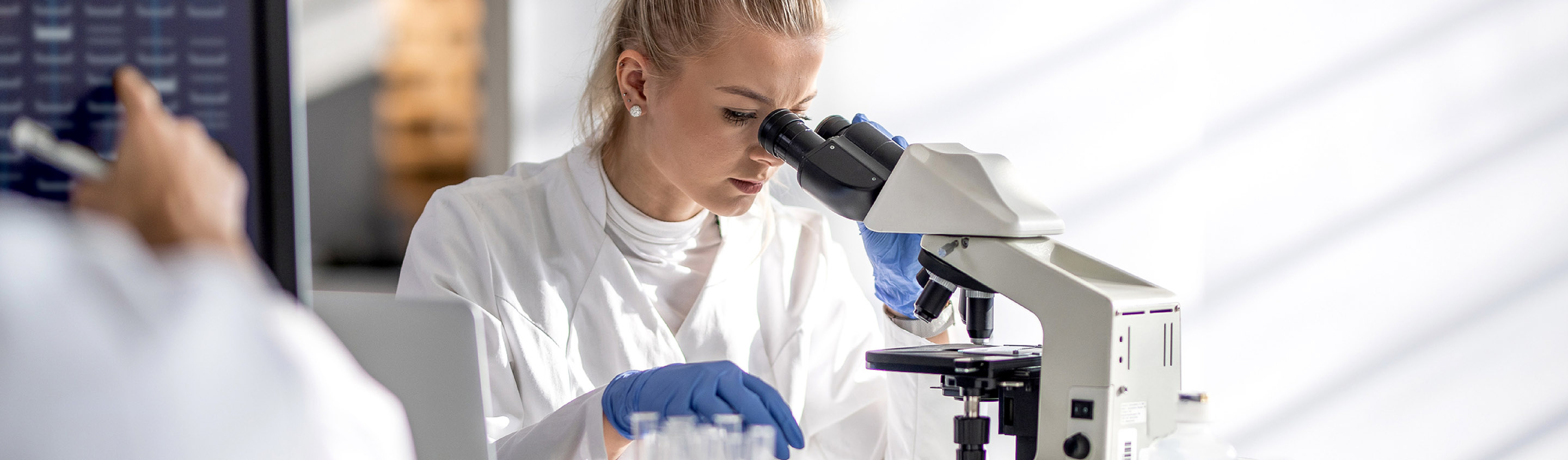 Scientist in lab coat and gloves examining a sample under a microscope
