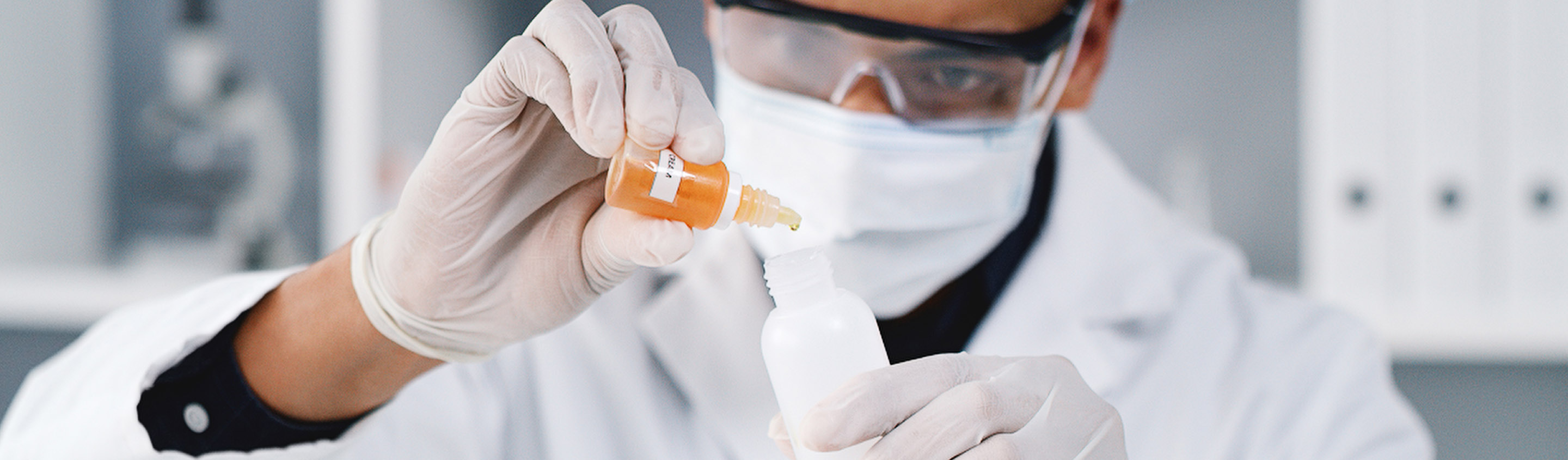 Scientist in mask and safety glasses inspecting a small vial of orange liquid