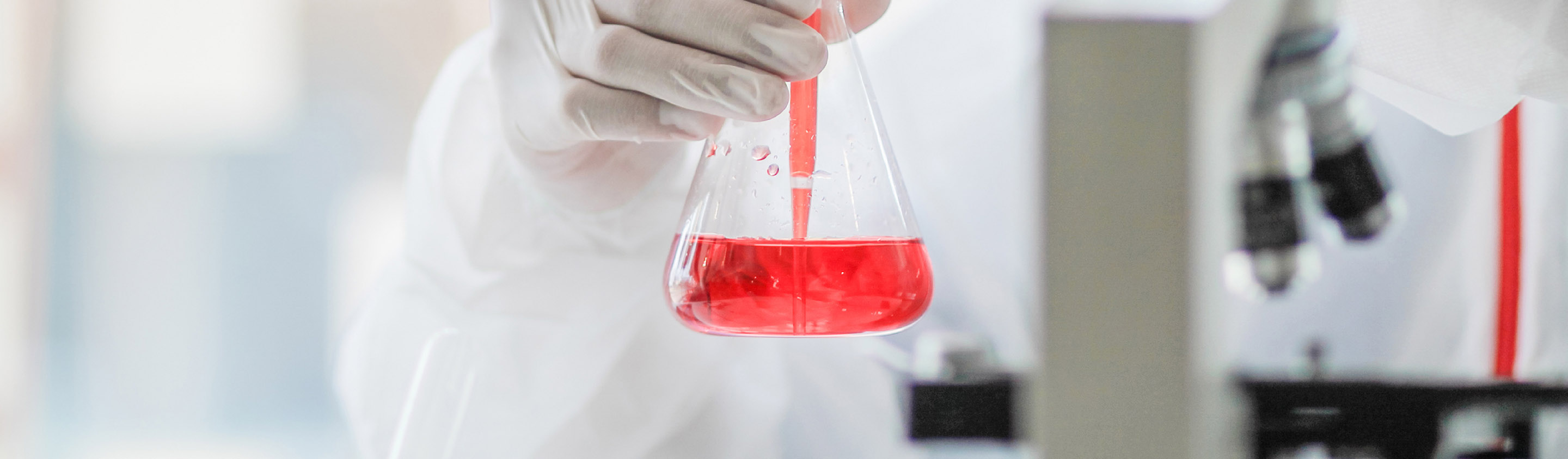 Scientist holding an Erlenmeyer flask of red liquid beside a microscope