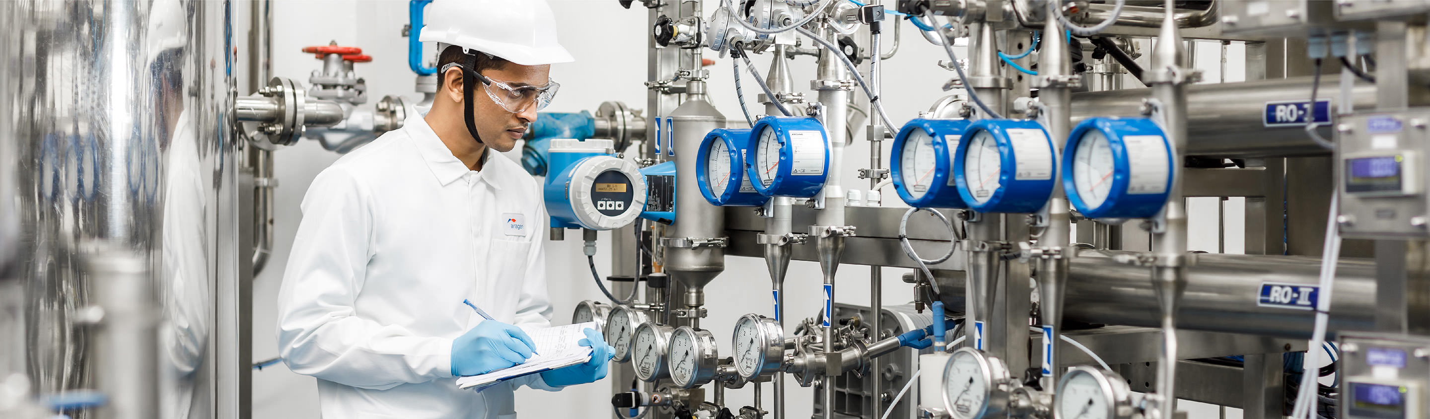 Aragen operator in hard hat and lab coat inspecting flow meters on process piping in a performance chemicals plant