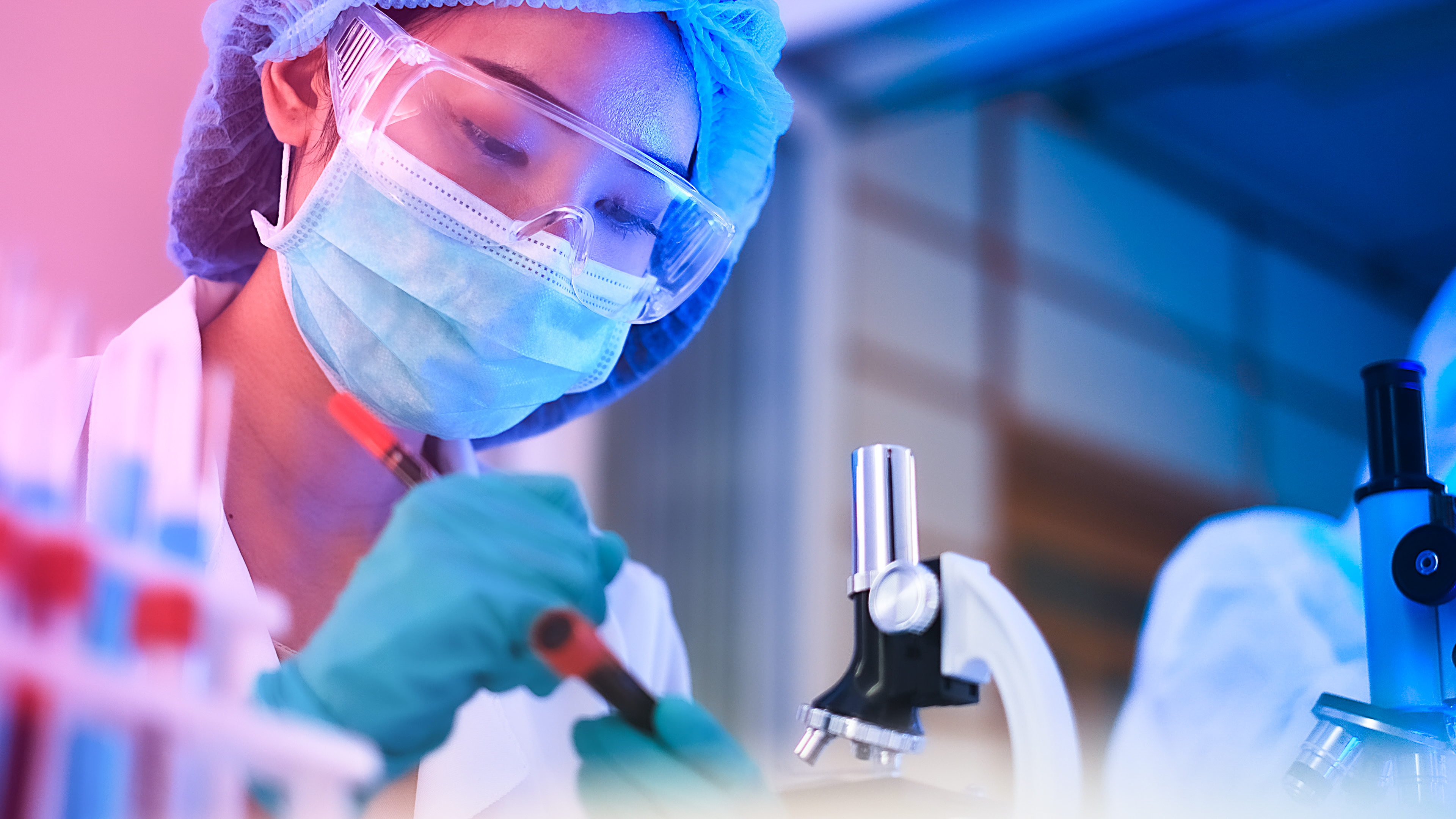 Scientist in cap, mask and goggles pipetting a red sample beside a microscope under blue and pink lighting