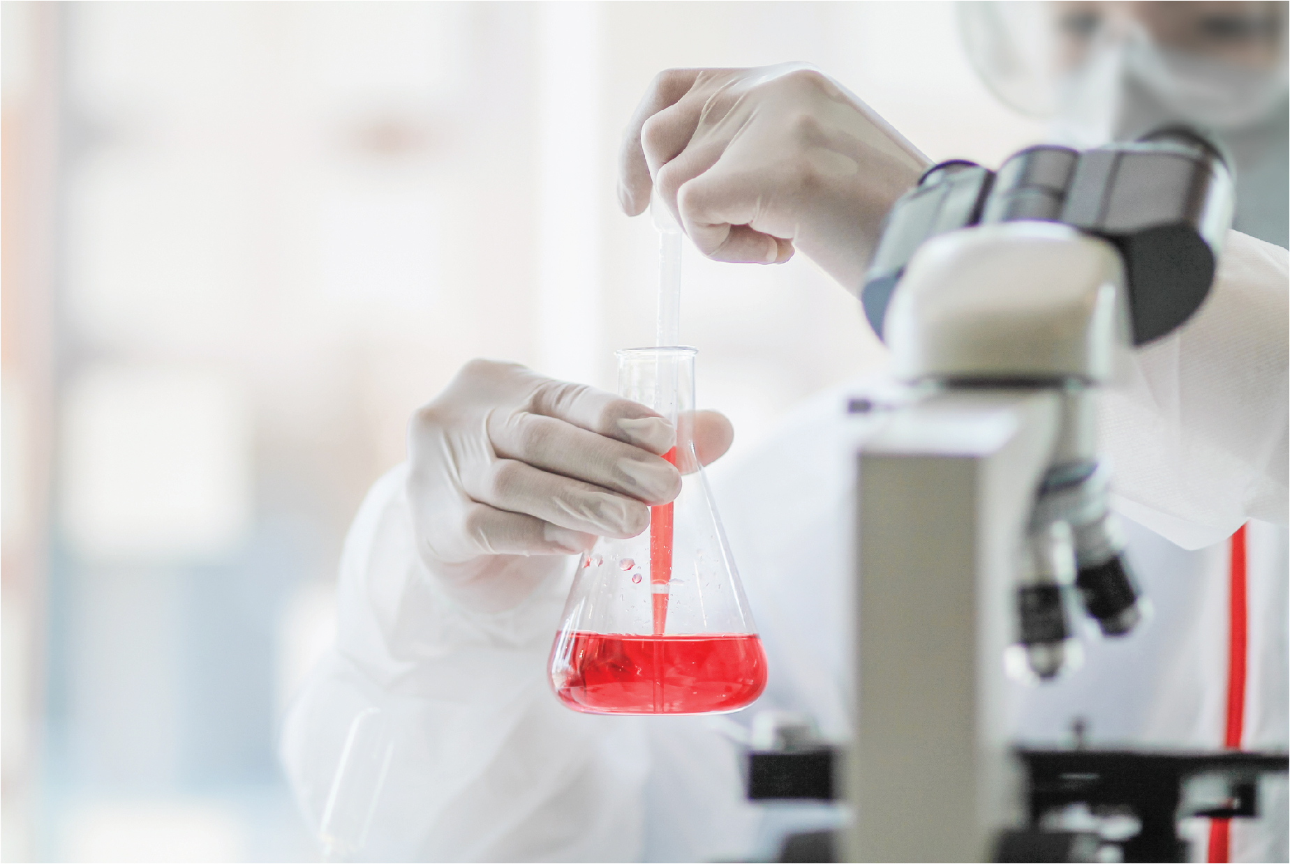 Gloved hand pipetting red liquid into an Erlenmeyer flask beside a microscope