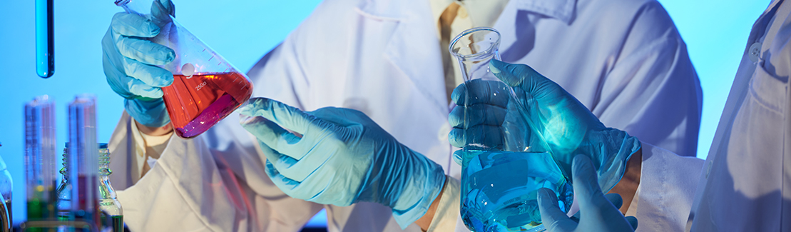 Two chemists in lab coats and gloves handling flasks of red and blue solution