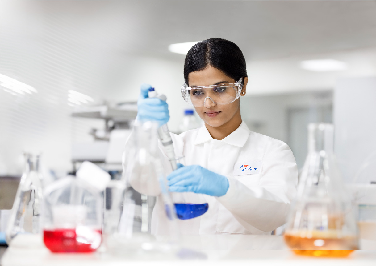 Aragen scientist in safety glasses and gloves working with flasks of coloured solutions at a laboratory bench