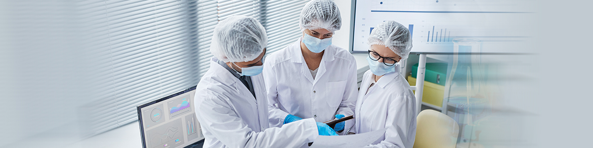 Three Aragen scientists in caps and masks discussing at a computer in a process validation lab