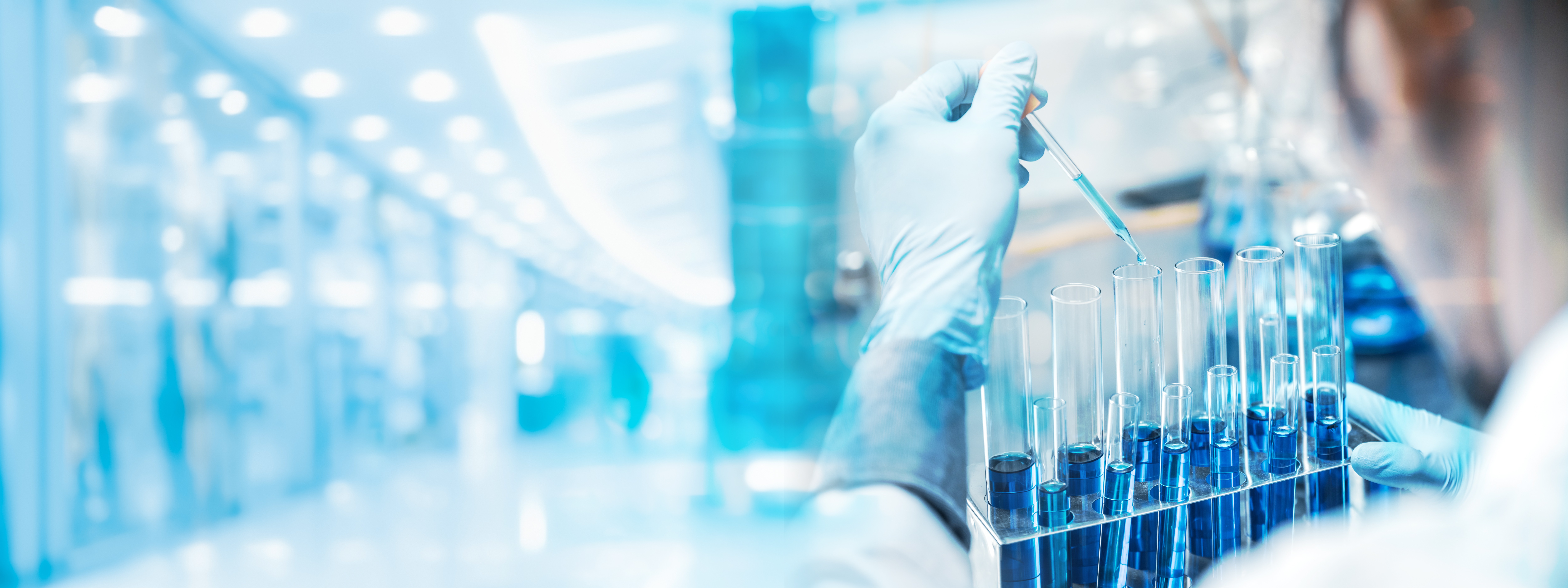 Gloved scientist pipetting into a rack of test tubes under blue lighting, representing hit-to-lead