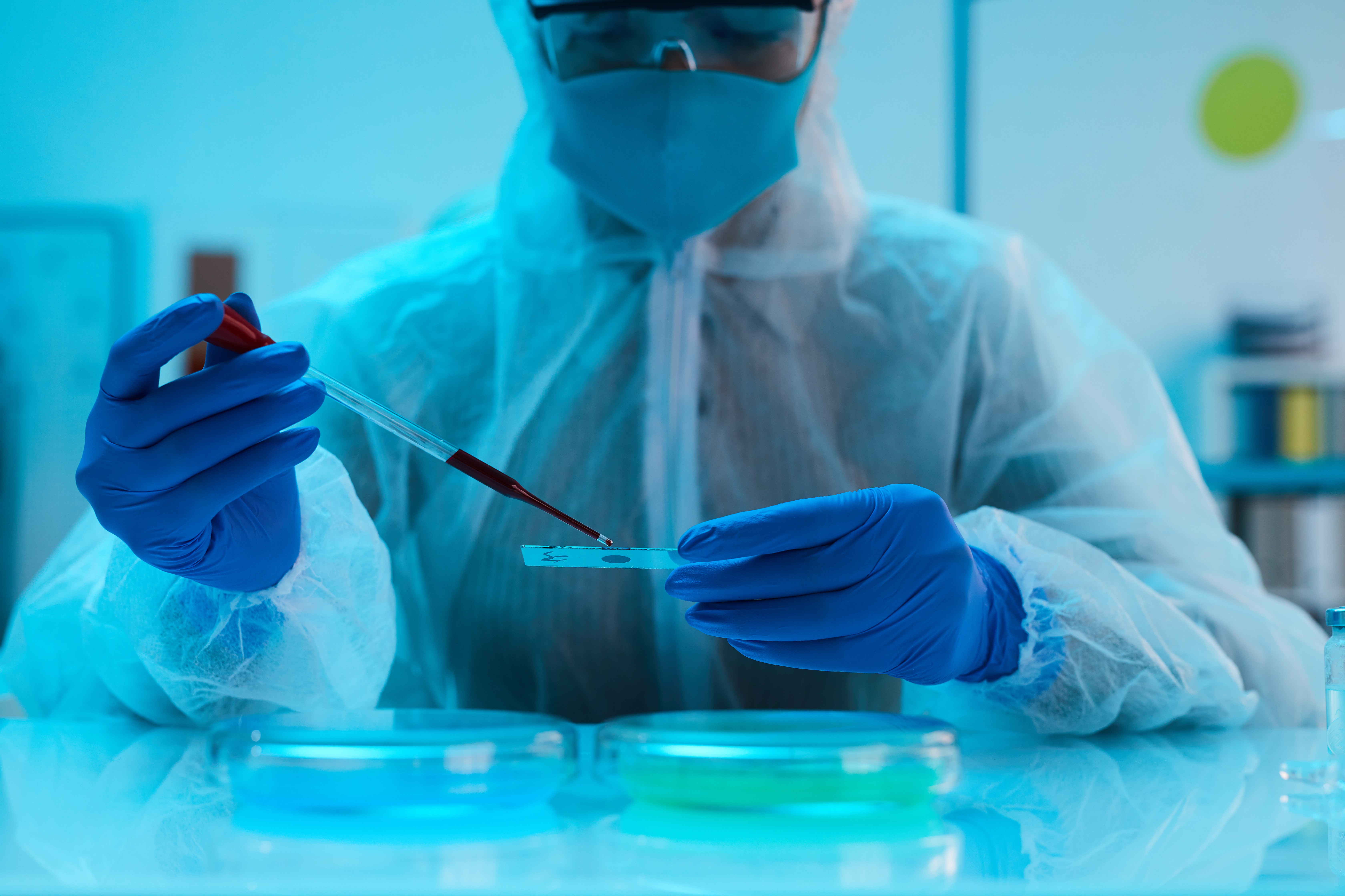 Scientist in blue protective suit and goggles pipetting into petri dishes, representing in vitro assay development