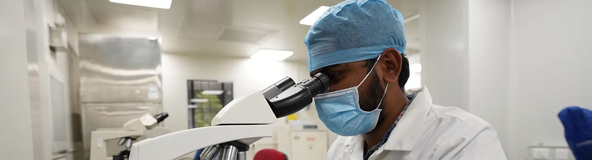 Aragen scientist in cap and mask looking through a microscope in a bioanalytical laboratory