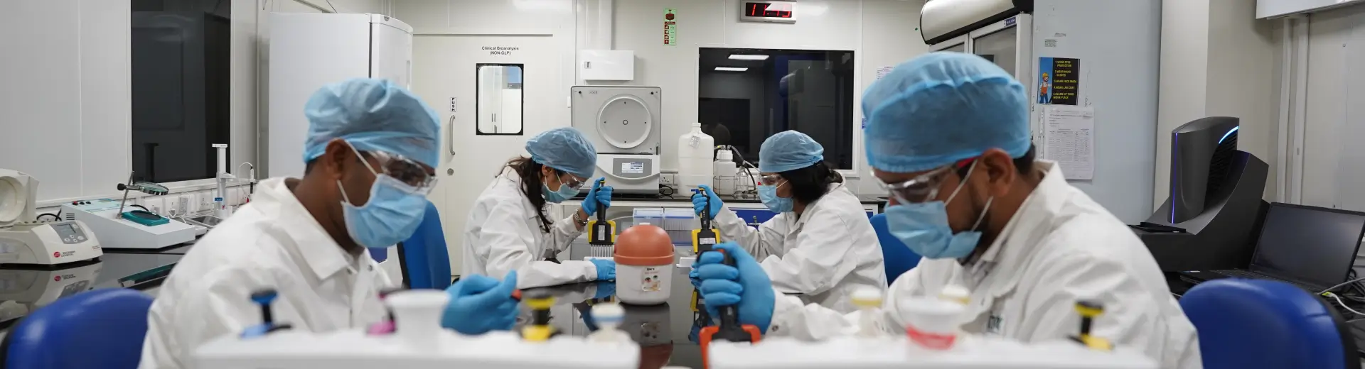 Aragen scientists in caps and masks working together in a laboratory, facility and compliance banner