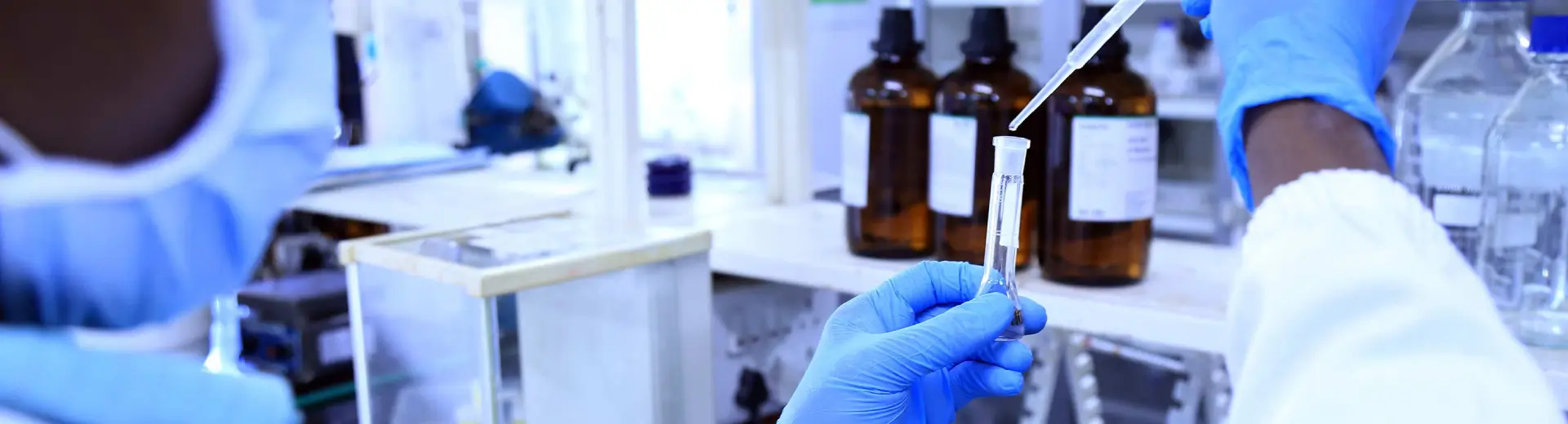 Gloved scientist pipetting from amber reagent bottles, quality systems banner